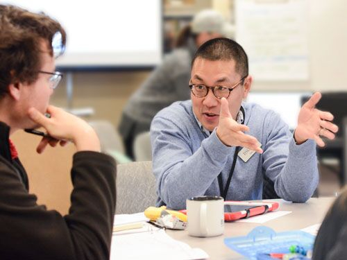 Man with glasses sitting at table talking to another person.