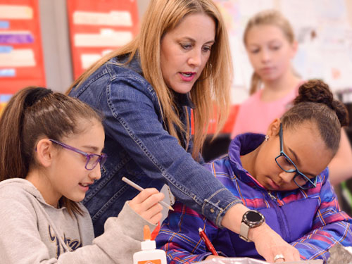 teacher with long hair and denim jacket pointing at something on a desk with three female fifth-grade students