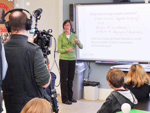 Female teacher in green sweater talking to her young students with two men filming her teach.